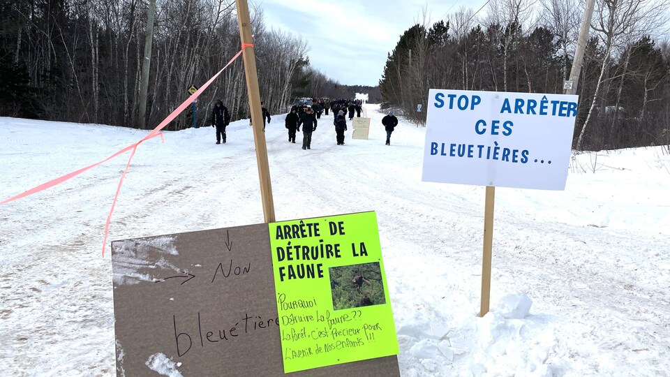 Des gens marchent dans la neige en direction d'une affiche plantée dans le sol sur laquelle est écrit « Stop Arrêter ces bleuetières...»