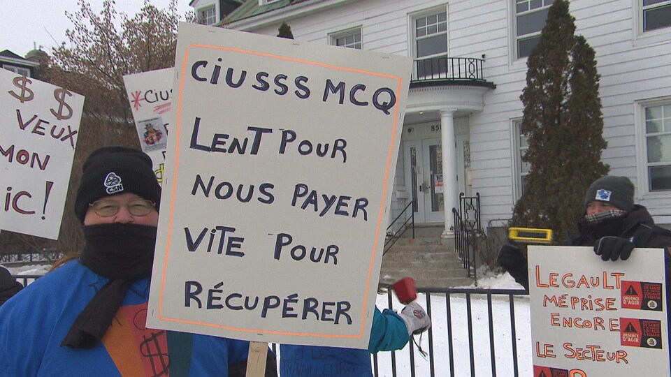 Une dame qui tient une pancarte sur laquelle on peut lire : CIUSSS MCQ lent pour nous payer vite pour récupérer.