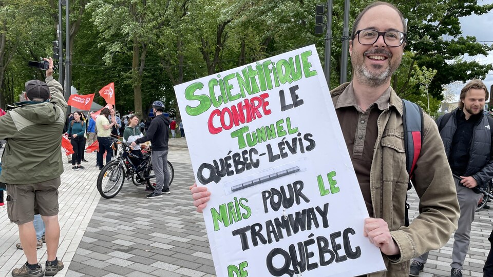 Un homme présente une pancarte à la caméra sur laquelle on peut lire ''Scientifique contre le tunnel Québec-Lévis mais pour le tramway de Québec''.