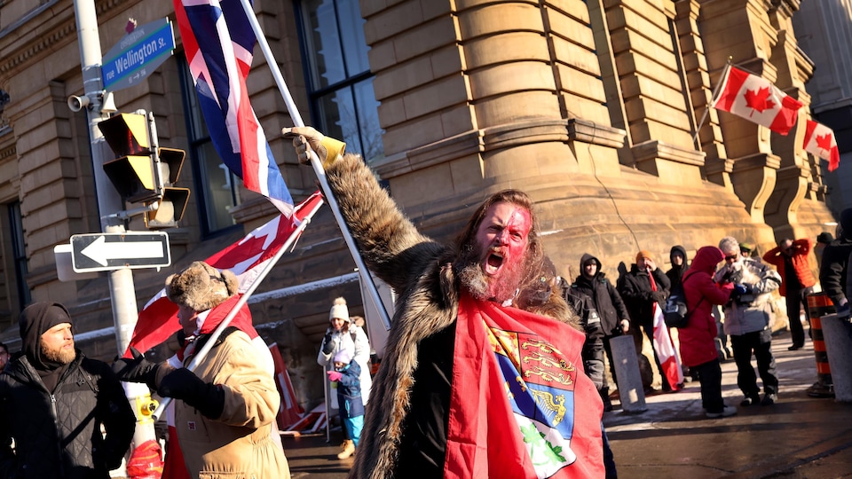 Un homme barbu et maquillé brandit différents drapeaux en criant.