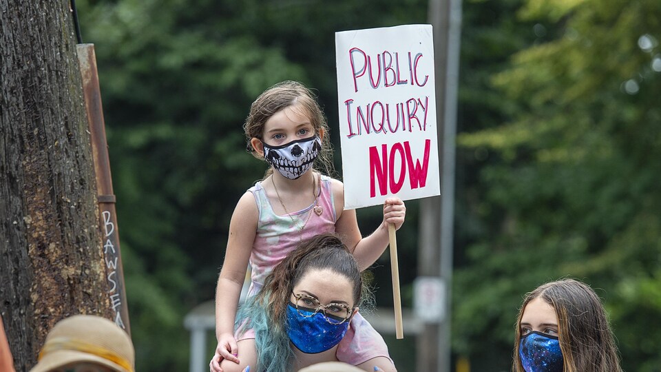 Une petite fille tient une pancarte et elle est assise sur les épaules d'une femme adulte. Toutes les personnes portent un masque contre le coronavirus.