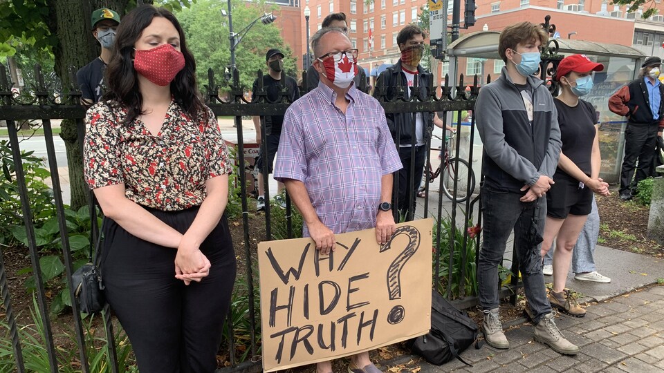Des personnes en rang manifestent silencieusement. Ils sont debout et on le visage couvert de masques contre le coronavirus.
