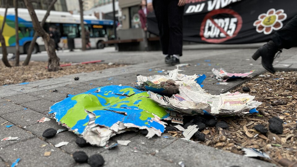 Des morceaux de la Terre en carton et de charbon éparpillés au sol, tandis qu'on voit à l'arrière les jambes d'une personne en pantalon et une banderole portant un signe «non à la COP15».