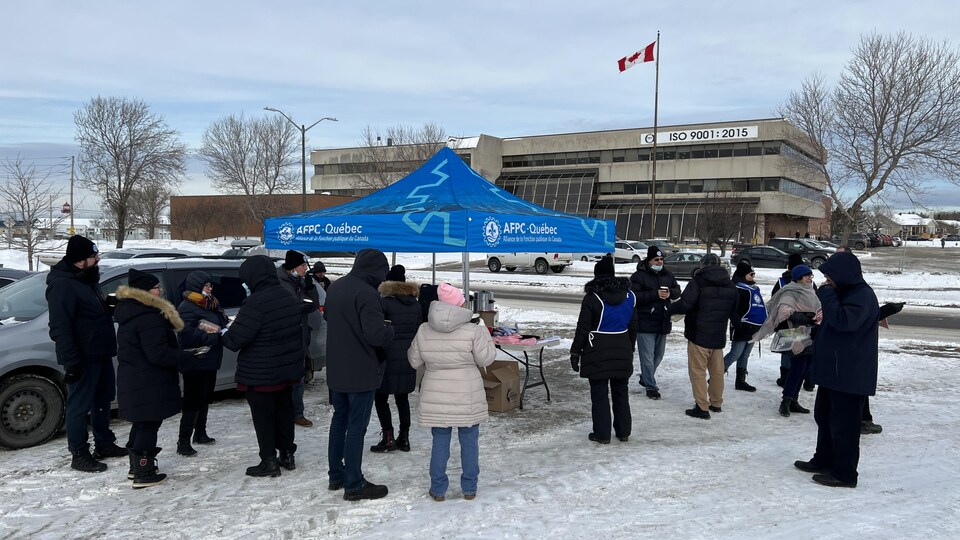 Un vingtaine de personnes prennent un café sous un chapiteau bleu en hiver. Devant elles, un drapeau canadien flotte face à un gros bâtiment.