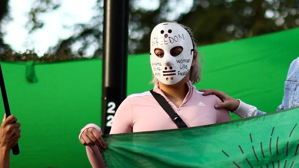 Au milieu d'une manifestation publique en appui au mouvement de contestation en Iran, une personne porte un masque de Jason Voorhees sur lequel les mots « Woman Life Freedom » sont écrits.