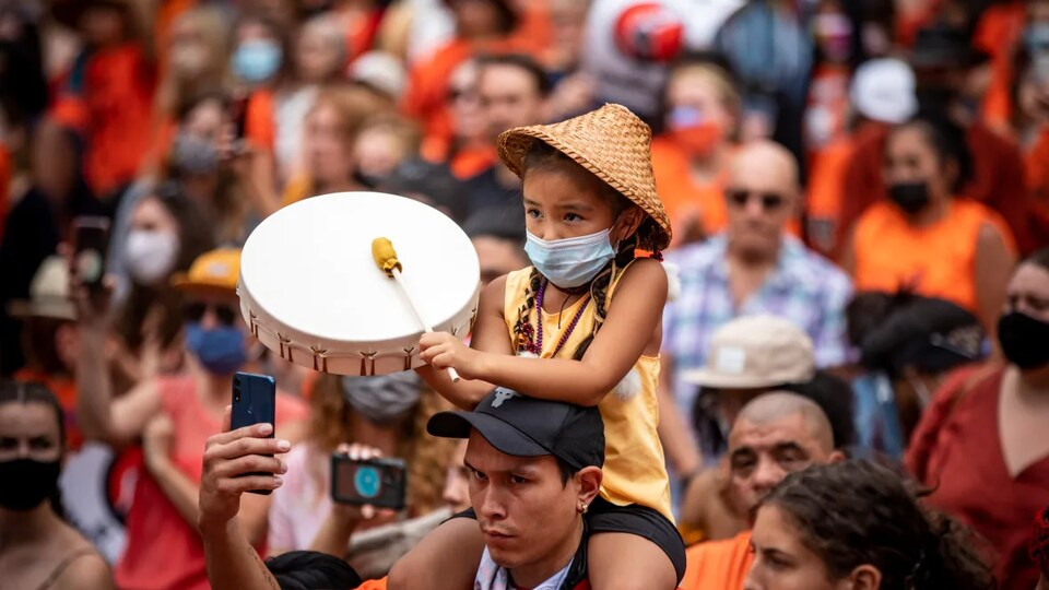 Une foule habillée avec des vêtements orange avec un enfant qui joue du tambour sur les épaules d'un adulte.