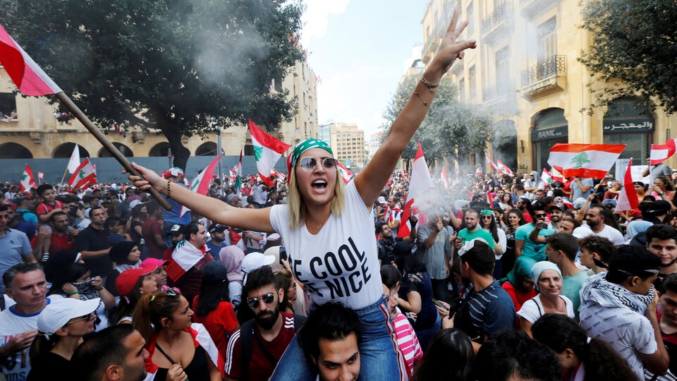 Une femme tient le drapeau du Liban et est assise sur les épaules d'un homme. Tout autour d'elle, une foule de manifestants. 