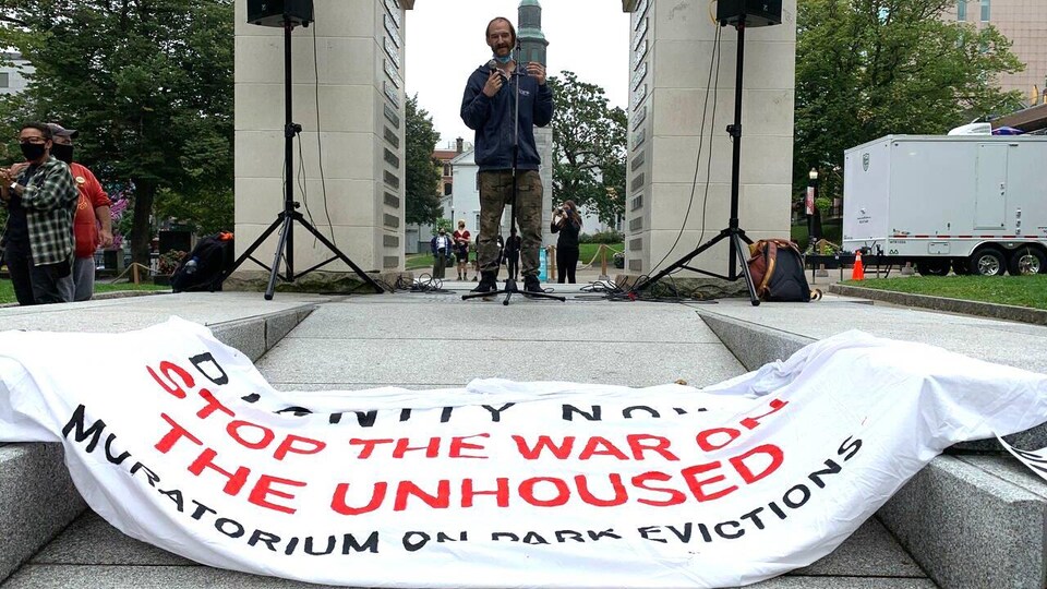 Un homme prend la parole en public. Sur le sol devant lui se trouve une banderole demandant que cesse la « guerre à ceux qui sont sans-abri ».