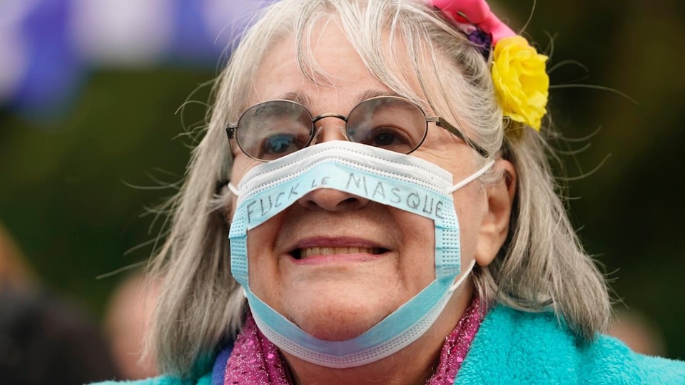 Une femme porte un masque qui est troué et sur lequel est écrit « fuck le masque ».                         