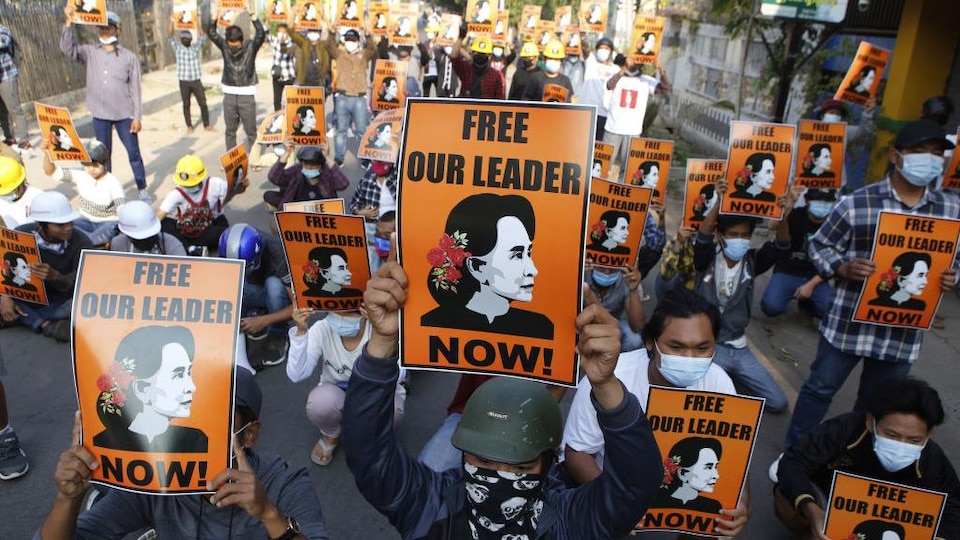 Des gens portant un masque tiennent des pancartes représentant le visage d'Aung San Suu Kyi, où l'on peut lire : « Libérez notre leader maintenant! »