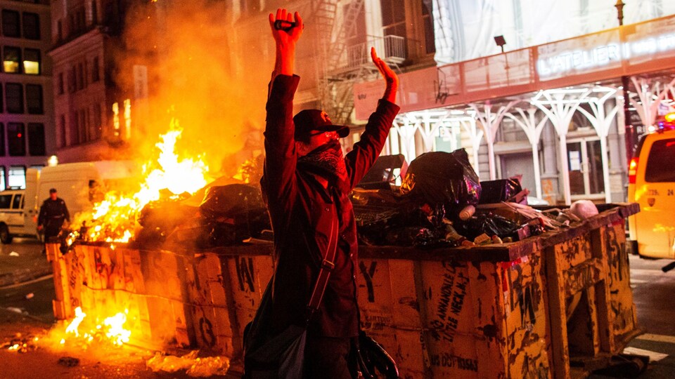Un manifestant, les bras dans les airs, devant un conteneur à ordures en feu