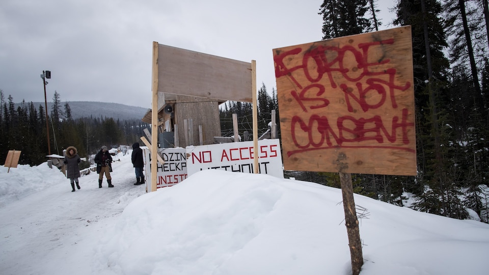 Plan serré d'une pancarte plantée dans la neige avec en arrière-plan des manifestants. Sur la pancarte il est écrit en anglais : il n'y a pas de consentement par la force.