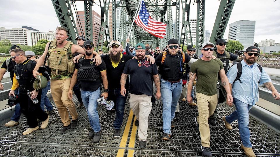 Des hommes traversent un pont à pied se tenant par les épaules et brandissant un drapeau américain. 