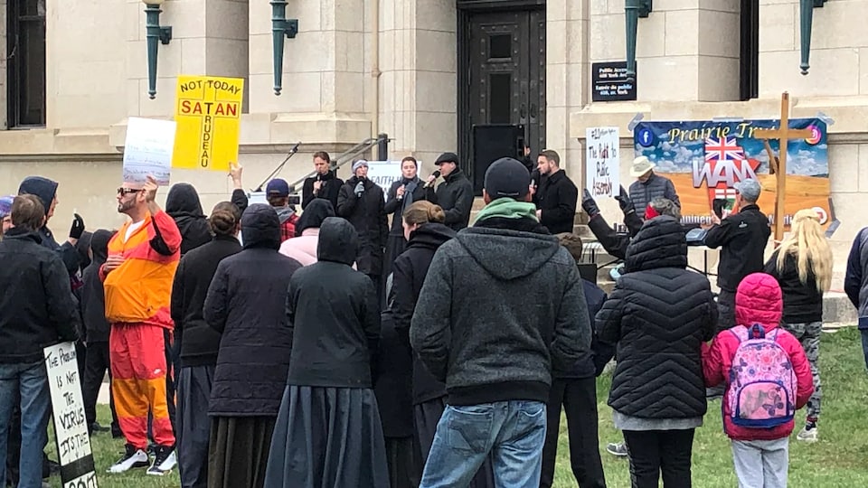 Une vingtaine de personnes rassemblées devant un bâtiment. Cinq personnes se tiennent devant les autres avec un micro. Des manifestants brandissent des affiches ainsi que des croix.