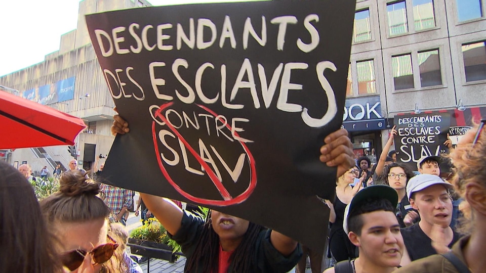 Une manifestante tient une pancarte sur laquelle on peut lire « Descendants des esclaves contre Slav ».