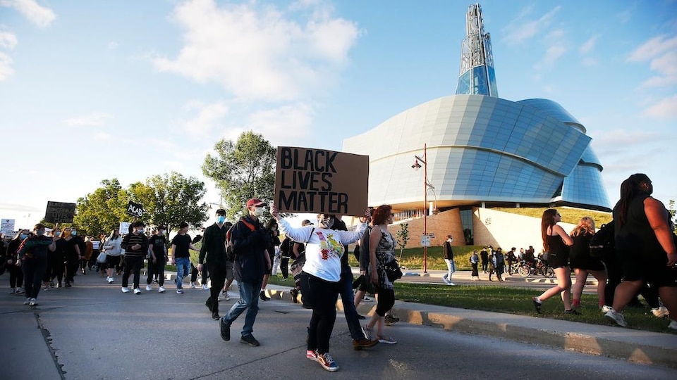 Des personnes marchent dans une rue en tenant des affiches, devant le Musée canadien pour les droits de la personne.