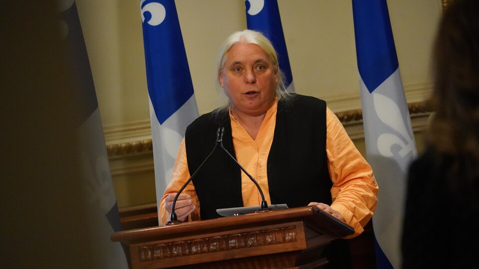 A woman speaks into the microphone in front of three fleur-de-lis.