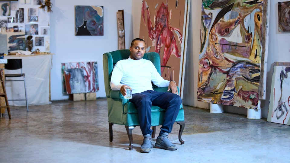 L'homme est assis sur un fauteuil et entouré de peintures.