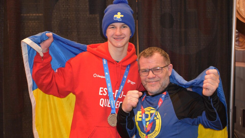 Un jeune homme avec la médaille d'or dans le cou, une tuque des Jeux du Québec sur la tête et le drapeau des Jeux sur les épaules, tout sourire, montrant les poings.