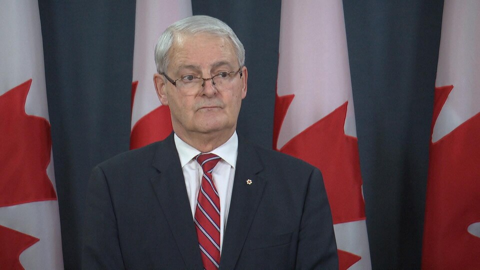 Marc Garneau, lors d'une conférence de presse devant des drapeaux canadiens.