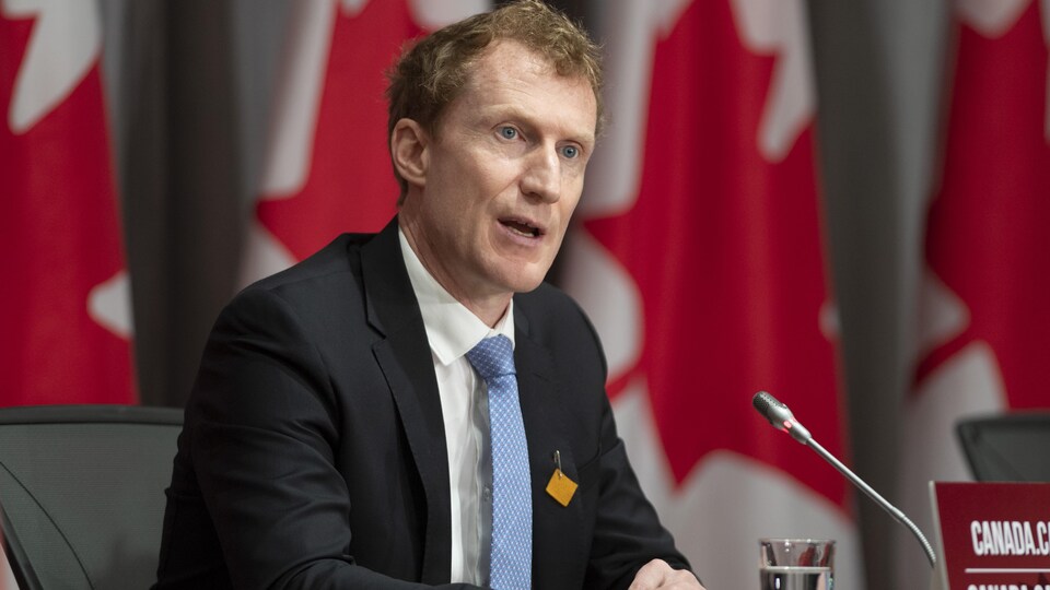 Marc Miller en conférence de presse devant plusieurs drapeaux du Canada.