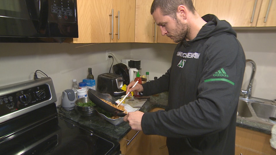 Un homme cuisine un repas avec des légumineuses devant sa cuisinière chez lui. Il porte un chandail à capuchon des Roughriders de la Saskatchewan