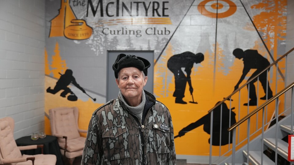 Un homme se tenant devant une enseigne intérieure du Club de curling McInyre.