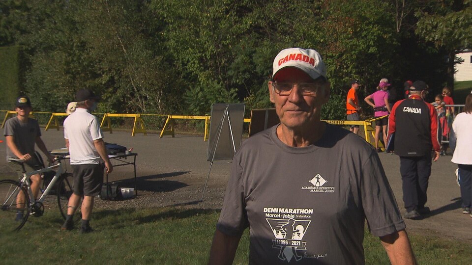 Marcel Jobin prend la pose en bordure du parcours du Demi-marathon où se trouvent quelques participants et des organisateurs.