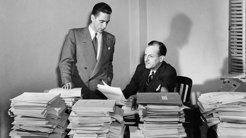 Dans un bureau, à Radio-Canada, le secrétaire du Concours dramatique de Radio-Canada, Jean-Jules Trudeau (debout), et le directeur du réseau français de Radio-Canada, Marcel Ouimet, assis derrière une pile de textes reçus dans le cadre du concours.