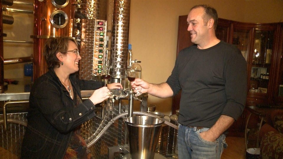 Un couple souriant qui tient des verres contenant un alcool sont debout devant un alambic de fer.