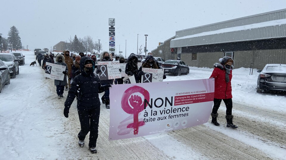 Deux femmes transportant une affiche sur laquelle est écrite l'inscription «Non à la violence faite aux femmes» marchent à la tête d'un groupe dans une rue d'Amos, en hiver, sous une faible neige.