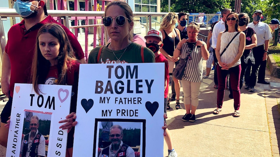 Les proches de victimes de la tuerie de Portapique marchant dans la rue. Une femme tient une affiche lisant Tom Bagley - mon père - ma fierté. Une jeune tient quant à elle une affiche lisant, Tom - grand-père - on s'ennuie.