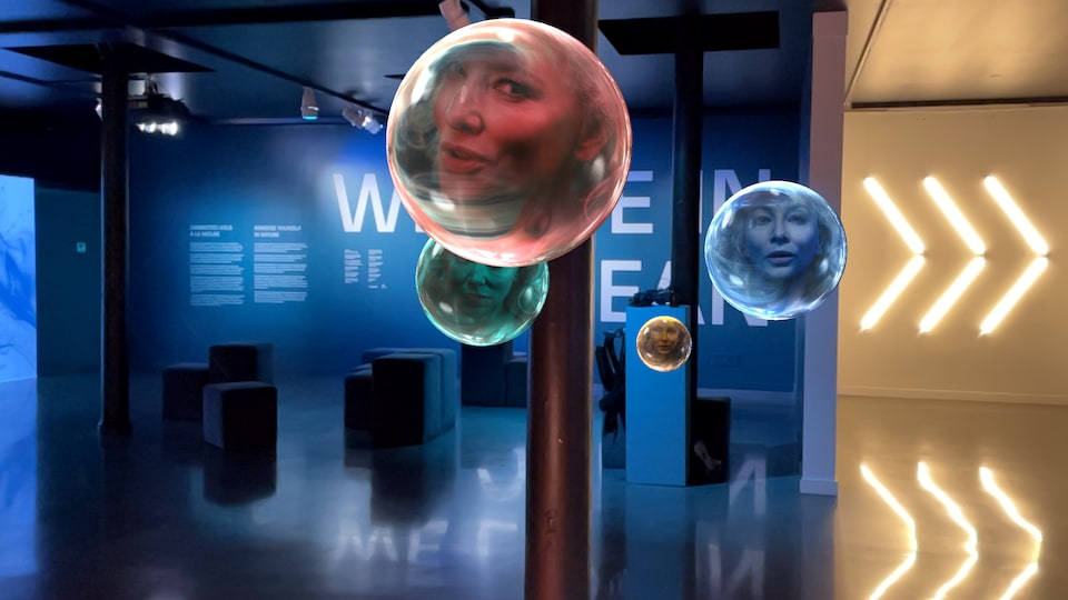Dans une salle d'exposition où des mots sont projetés sur un mur, on voit s'élever ce qui ressemble à des bulles qui flottent, dans lesquelles on voit le visage d'une personne.