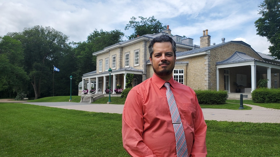 Un homme en chemise, cravate nouée autour du cou, pose dans le jardin d'une belle maison.