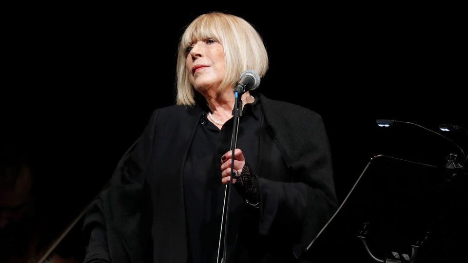 Marianne Faithfull, toute vêtue de noir, est sur scène et tient le pied d'un mi.