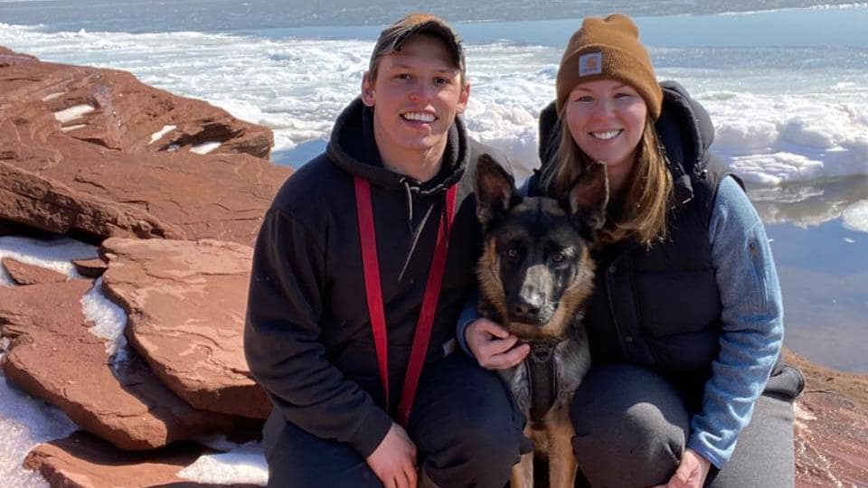 Marianne Tremblay-Abel et son conjoint Timothy James Markland posent pour la caméra avec un chien, à côté d'une falaise et de la mer gelée durant l'hiver.