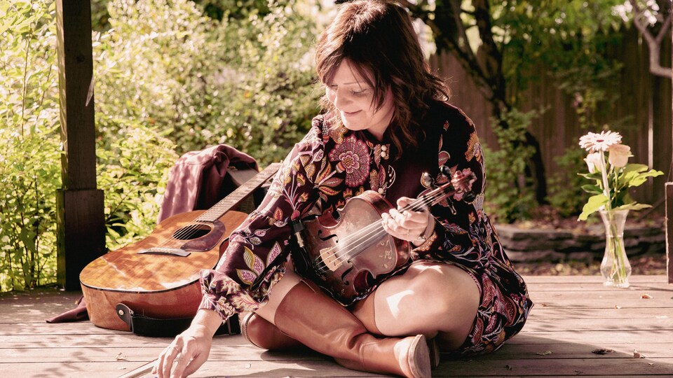Une femme assise en tailleur à l'extérieur, un violon à la main. 