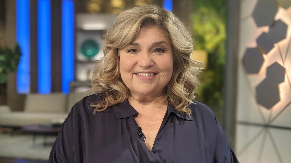 Une femme blonde, debout dans un décor de télévision, porte une chemise noire et sourit.