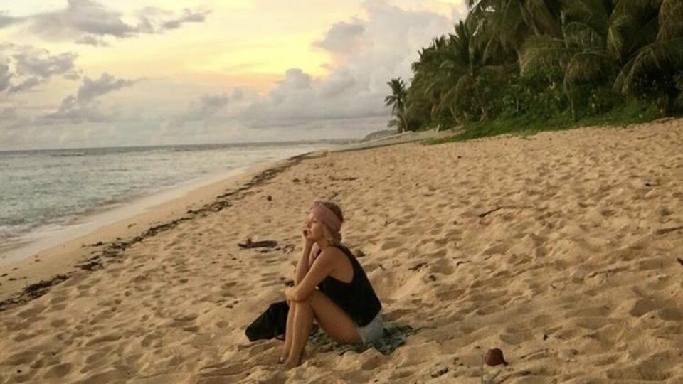 La femme est assise dans le sable fin. Au loin, on voit un coucher de soleil, la mer et des palmiers.