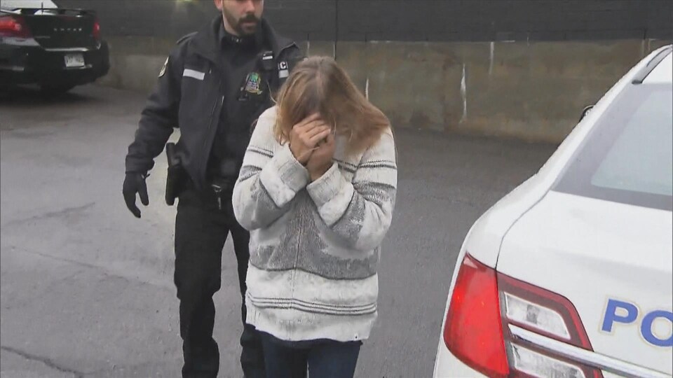 Marie-Jeanne Côté, escortée par un policier, se cache le visage avec les mains.