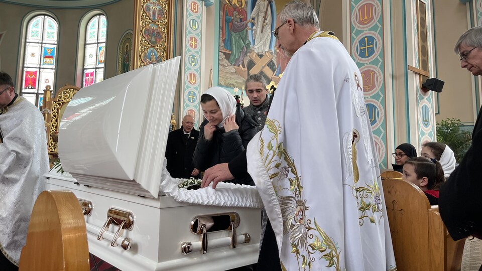 Les parents de Mariia, accompagnés du célébrant, se penchent sur le cercueil de l'enfant. La mère a un regard aimant.