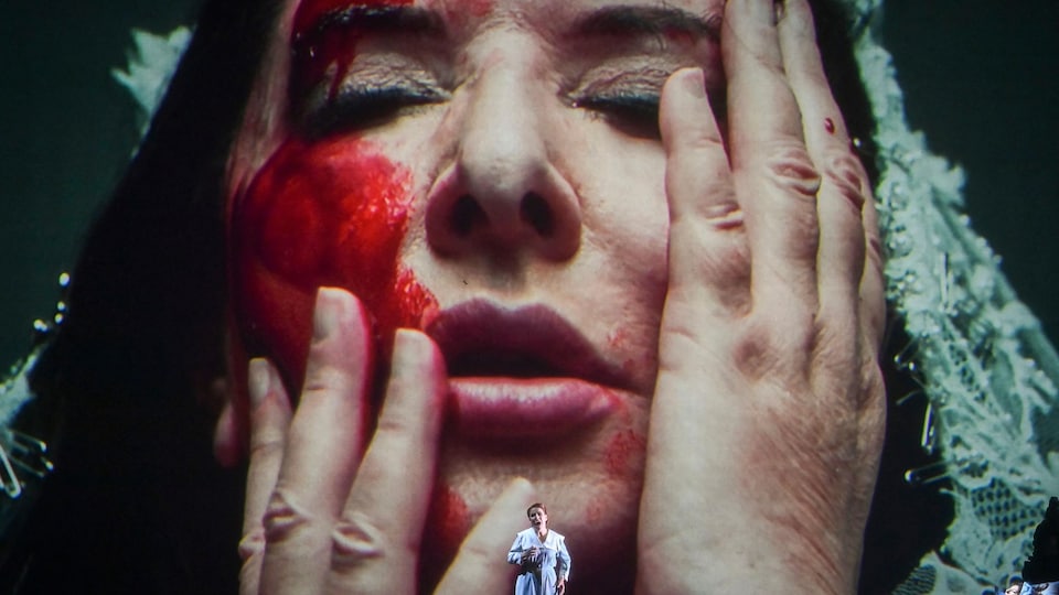 Une artiste donne une performance sur scène devant un écran géant qui projette son propre visage.