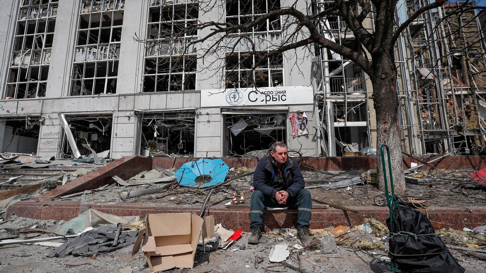 Un homme est assis devant un immeuble dont toutes les vitres ont volé en éclats. Des débris jonchent le sol et un arbre est carbonisé tout près.