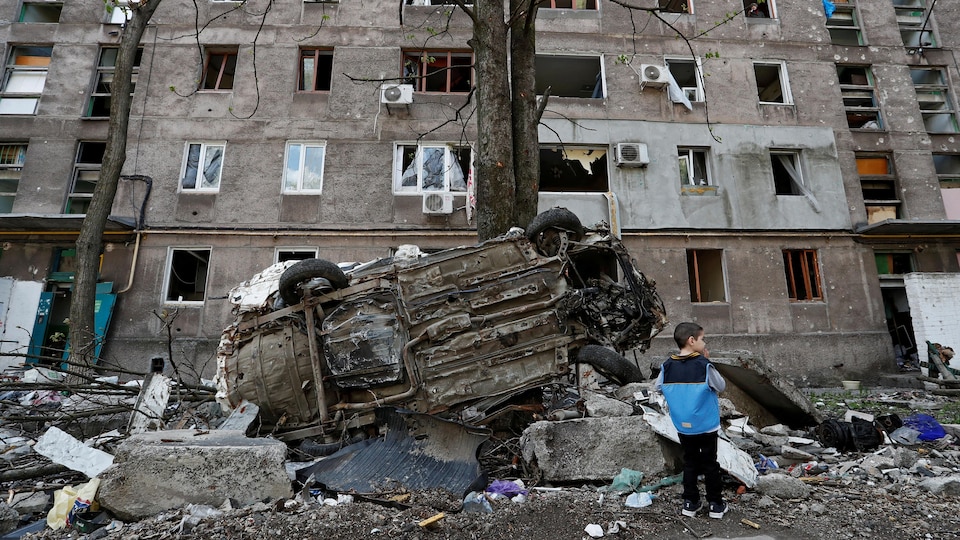 Un garçon, de dos, près d'un véhicule renversé devant un immeuble endommagé.