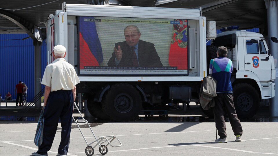 Two men stared at a screen installed on the bed of a truck.  It features Russian President Vladimir Putin. 