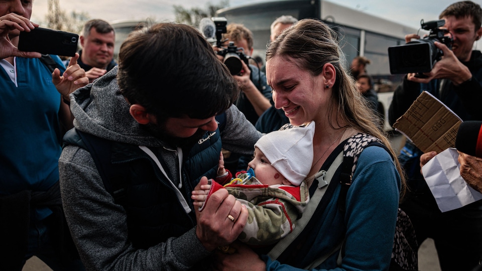 Un homme a la main gauche sur l'épaule d'une femme et serre de la main gauche le bras d'un bébé qu'elle tient sur son ventre. 