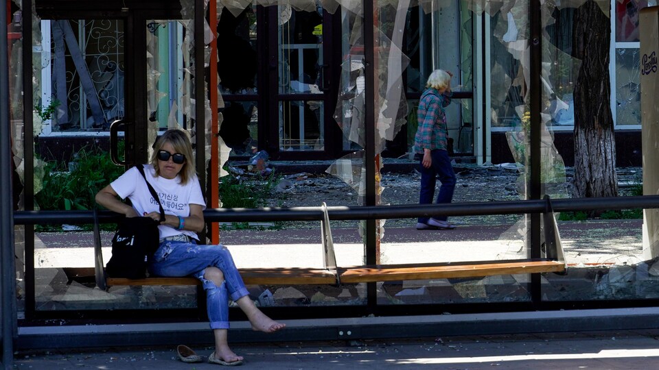 A woman was sitting in a bus shelter with all the windows broken. 