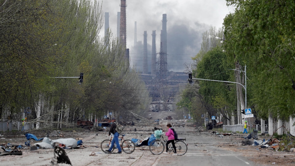 Des gens marchent à côté de leur vélo sur une route menant à une usine d'où s'échappe de la fumée. 
