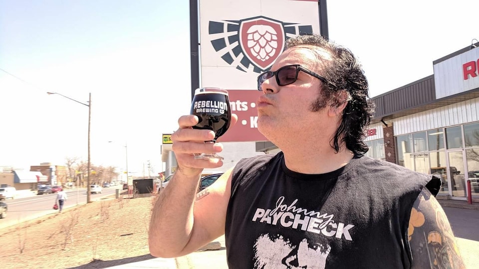 Un homme s'apprête à boire une bière brune de Rebellion Brewing devant les locaux de la brasserie à Regina lors d'une journée ensoleillée.