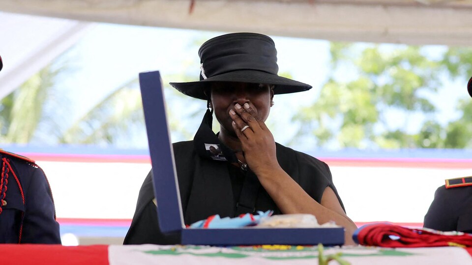 Martine Moïse, habillée en noir, porte une main à sa bouche devant un cercueil recouvert d'un drapeau haïtien.